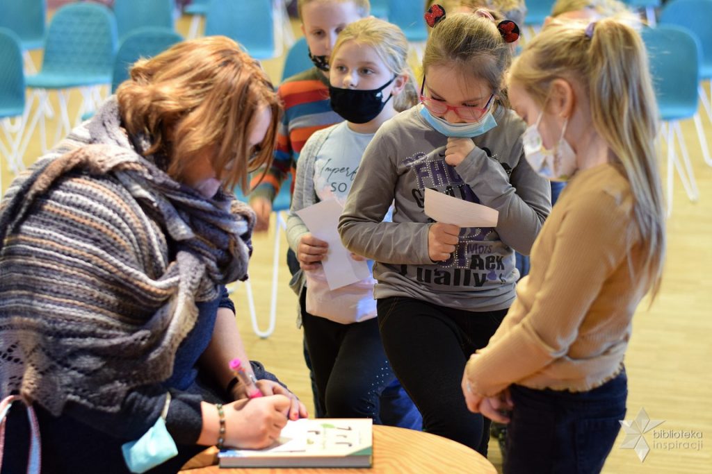Pani Justyna Bednarek piszę dedykację dla uczestników spotkania, w tle kolejka dzieci do autografu