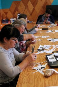 Uczestnicy Klubu Seniora siedzą przy dużym stole podczas zajęć artystycznych i wycinają wzory w papierowych serwetkach