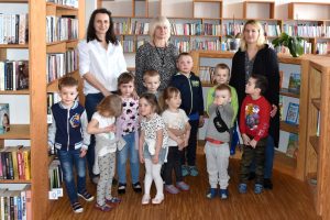 Dzieci, Panie z przedszkola oraz Dyrektor GBP w Wyrykach Hanna Czelej stoją między regałami książek w przestrzeni biblioteki