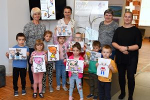 Dzieci trzymając w dłoniach swoje ilustracje o Kici Koci stoją wraz z wychowawczyniami oraz Hanną Czelej – Dyrektor GBP w Wyrykach pozując do zdjęcia, w tle plakat projektu
