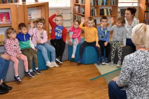 Dzieci wraz z wychowawczyniami siedzą w przestrzeni biblioteki na pufach w półokręgu, większość dzieci podnosi rękę do góry