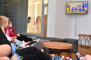 Dzieci siedzą w przestrzeni biblioteki na pufach i oglądają krótką animację o Kici Kocia na telewizorze zawieszonym na ścianie