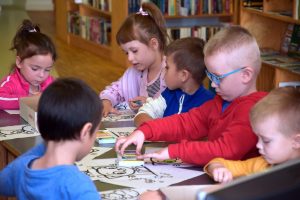 Dzieci stoją przy ladzie bibliotecznej, kolorują ilustracje o Kici Koci, w tle regały z książkami