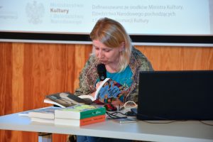Na scenie Pani Małgorzata Karolina Piekarska siedzi przy biurku czyta fragment swojej książki. W tle plakat spotkania wyświetlony na ekranie ściennym