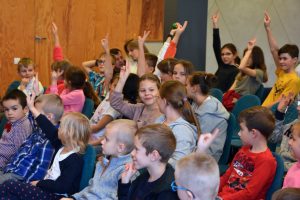 Dzieci podczas spotkania autorskiego siedzą na krzesłach przed sceną, większość z nich zgłasza się do odpowiedzi poprzez podniesienie ręki do góry