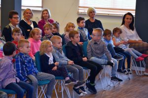 Dzieci podczas spotkania autorskiego siedzą na krzesłach, przed dziećmi trzymając w dłoni mikrofon stoi Pan Daniel de Latour