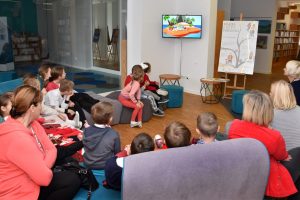 W przestrzeni biblioteki w półokręgu siedzą dzieci wraz z nauczycielami, oglądają bajkę o emocjach na zawieszonym na ścianie TV, w tle plakat wydarzenia oraz półki z książkami