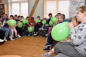 W przestrzeni biblioteki w półokręgu siedzą dzieci pompując zielone balony