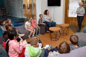 W przestrzeni biblioteki w półokręgu siedzą dzieci wraz z nauczycielem, po przeciwnej stronie stoi Hanna Czelej – Dyrektor GBP Wyryki