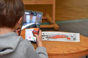 Na pierwszym planie chłopiec trzyma tablet i za pomocą aplikacji ożywia i nadaje swojej kolorowance trójwymiarowe kształty, na drugim planie na stoliku leży kolorowanka