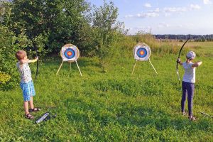 Po lewej i prawej stoją młodzi łucznicy, w rękach trzymają łuk oraz strzały wycelowane w tarce strzelnicze stojące przed nimi