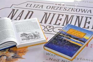 Na stole leży plakat wydarzenia, na nim dwie książki Elizy Orzeszkowej „Nad Niemnem”