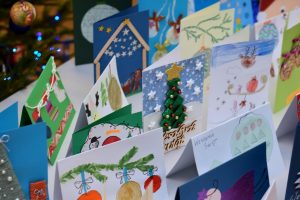 Na zdjęciu widać wiele kolorowych kartek świątecznych ustawionych na białym stole. Każda kartka jest unikalna, posiada różne wzory, kolory i tekstury. Kartki są różnych rozmiarów i kształtów, niektóre wykonane ręcznie z rysunkami choinek, bałwanów, domów pokrytych śniegiem i innych elementów świątecznych.