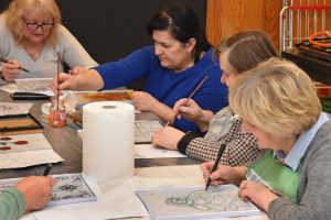 Członkinie Klubu Seniora „Aktywni” malują na arkuszach papieru. Każda osoba trzyma w ręku pędzel lub pisak i pracuje nad indywidualnym projektem.