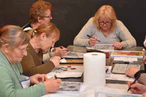 Członkinie Klubu Seniora „Aktywni” malują na arkuszach papieru. Każda osoba trzyma w ręku pędzel lub pisak i pracuje nad indywidualnym projektem.