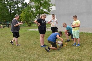 Na zdjęciu widzimy grupę uczestników wakacji z bibloteką, zaangażowanych w zabawę z obręczą hula-hop. Jedna osoba klęczy na trawie, pochylona do przodu z rękoma w obręczy, która leży płasko na ziemi. Cztery inne osoby stoją wokół klęczącej osoby. W tle drzewa, budynek szkoły i biblioteki.