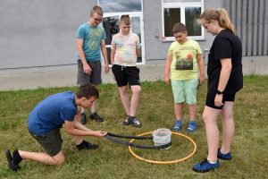 Na zdjęciu widać grupę sześciu osób na zewnątrz. Jeden z uczestników klęczy na ziemi, trzymając w ręku dętkę od roweru, którą wkłada do koła hula-hop leżącego na ziemi. Pozostałe pięć osób stoi i obserwuje. W tle widoczny jest budynek biblioteki.