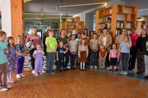 Na zdjęciu widać grupę ludzi w bibliotece. Biblioteka jest jasna i przestronna, z półkami pełnymi książek i nowoczesnym oświetleniem. Grupa składa się zarówno z dorosłych, jak i dzieci.