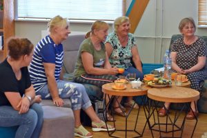 Zdjęcie w przestrzeni biblioteki przedstawiające uczestników spotkania DKK siedzących na kanapach wokół dwóch okrągłych, drewnianych stolików kawowych.