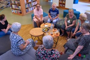Uczestnicy spotkania DKK rozmawiają w półkolu w bibliotece. Siedzą na szarych kanapach i pufach wokół dwóch okrągłych stolików kawowych. W tle widać regały pełne książek.
