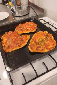 Duże zbliżenie na trzy małe, ręcznie robione pizze, leżące na czarnej blasze do pieczenia. Pizze są o różnych kształtach, z wyraźnie widocznym sosem pomidorowym, roztopionym serem i różnymi dodatkami.