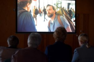 Kilka osób siedzi w rzędzie na krzesłach w bibliotece i wspólnie ogląda projekcję filmu na dużym ekranie. Na ekranie widać scenę z udziałem mężczyzny z plecakiem, rozmawiającego z inną osobą.