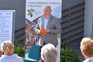 Wójt Gminy Wyryki Bernard Błaszczuk trzymający tomik poezji przemawia podczas wydarzenia „Narodowe Czytanie” przed Gminną Biblioteką Publiczną w Wyrkach. Obok widoczny jest plakat promujący wspólne czytanie poezji Jana Kochanowskiego oraz baner biblioteki. Przed mówcą siedzi kilku słuchaczy.
