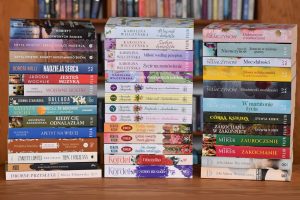 Jest to zdjęcie zrobione na wprost, przedstawiające trzy pionowe stosy książek w twardych i miękkich okładkach, stojące przed tłem z regałów wypełnionych innymi książkami.