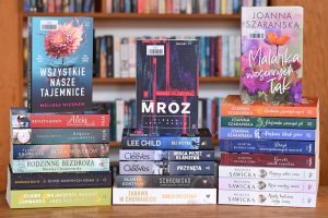 Jest to zdjęcie zrobione na wprost, przedstawiające trzy pionowe stosy książek w twardych i miękkich okładkach, stojące przed tłem z regałów wypełnionych innymi książkami.