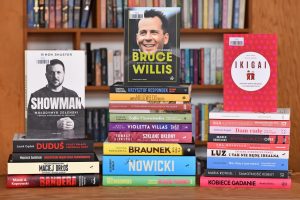 Jest to zdjęcie zrobione na wprost, przedstawiające trzy pionowe stosy książek w twardych i miękkich okładkach, stojące przed tłem z regałów wypełnionych innymi książkami.