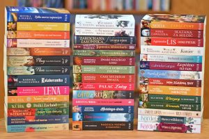 Jest to zdjęcie zrobione na wprost, przedstawiające trzy pionowe stosy książek w twardych i miękkich okładkach, stojące przed tłem z regałów wypełnionych innymi książkami.