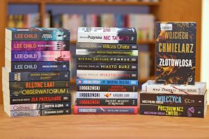 Jest to zdjęcie zrobione na wprost, przedstawiające trzy pionowe stosy książek w twardych i miękkich okładkach, stojące przed tłem z regałów wypełnionych innymi książkami.